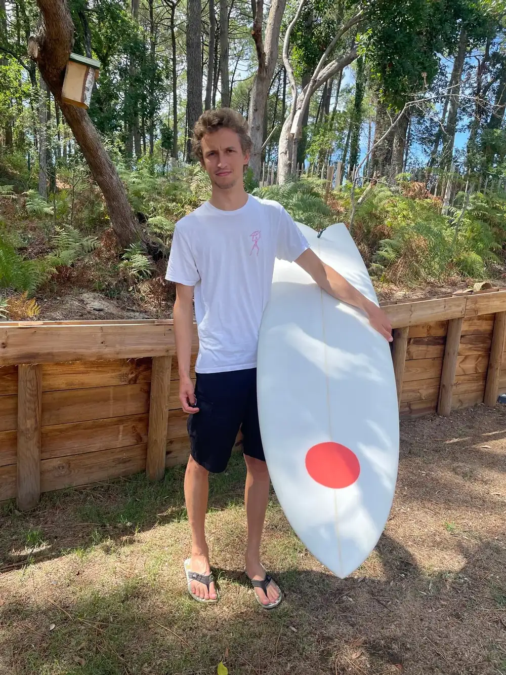 Achille souriant derrière sa planche twin fish Waxtoday, dans le jardin avant de partir surfer