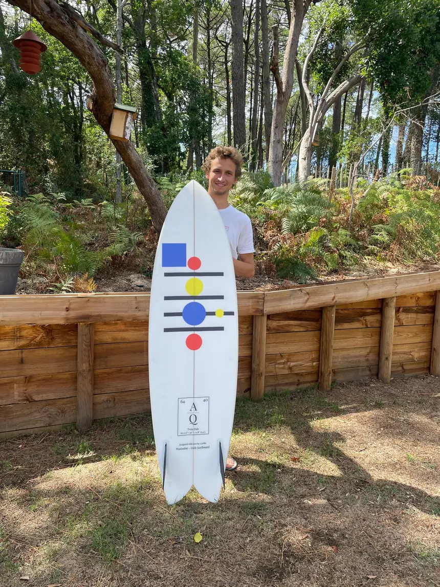Achille debout avec sa planche twin fish Waxtoday aux motifs Atomique